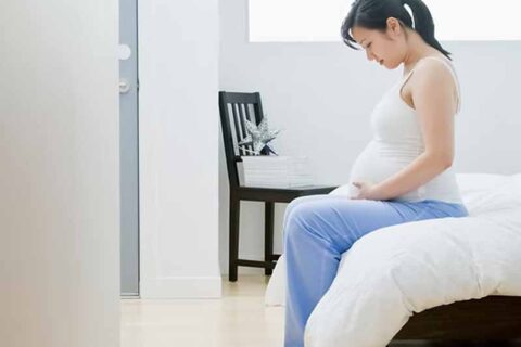 Pregnant person sitting on a bed in a bright, minimalistic room.