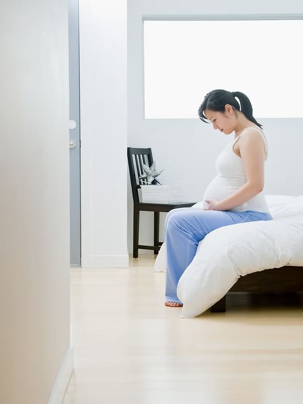 Pregnant person sitting on a bed in a bright, minimalistic room.