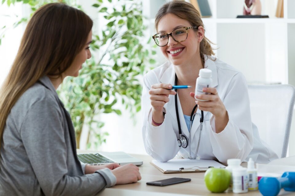 Person in a white coat holding a pill bottle and pen while speaking with another person seated at a desk in Lake Saint Louis, MO
